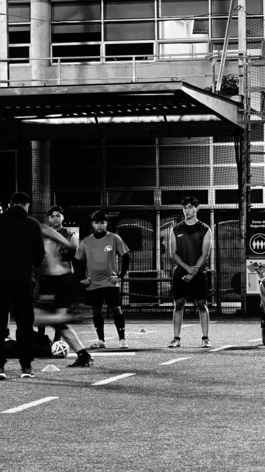 El equipo escucha al entrenador en una cancha iluminada de noche, en blanco y negro