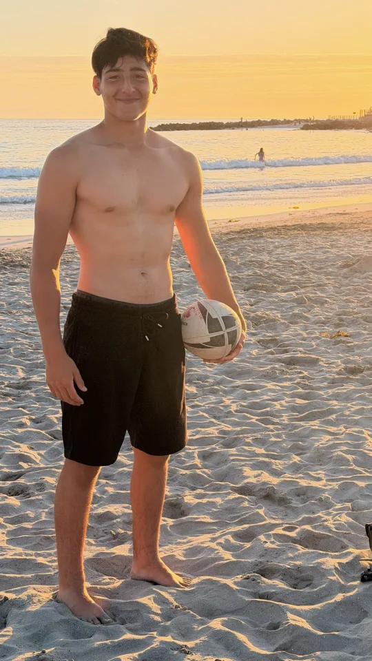 Joaquín de pie en la arena sosteniendo el balón de rugby, al atardecer
